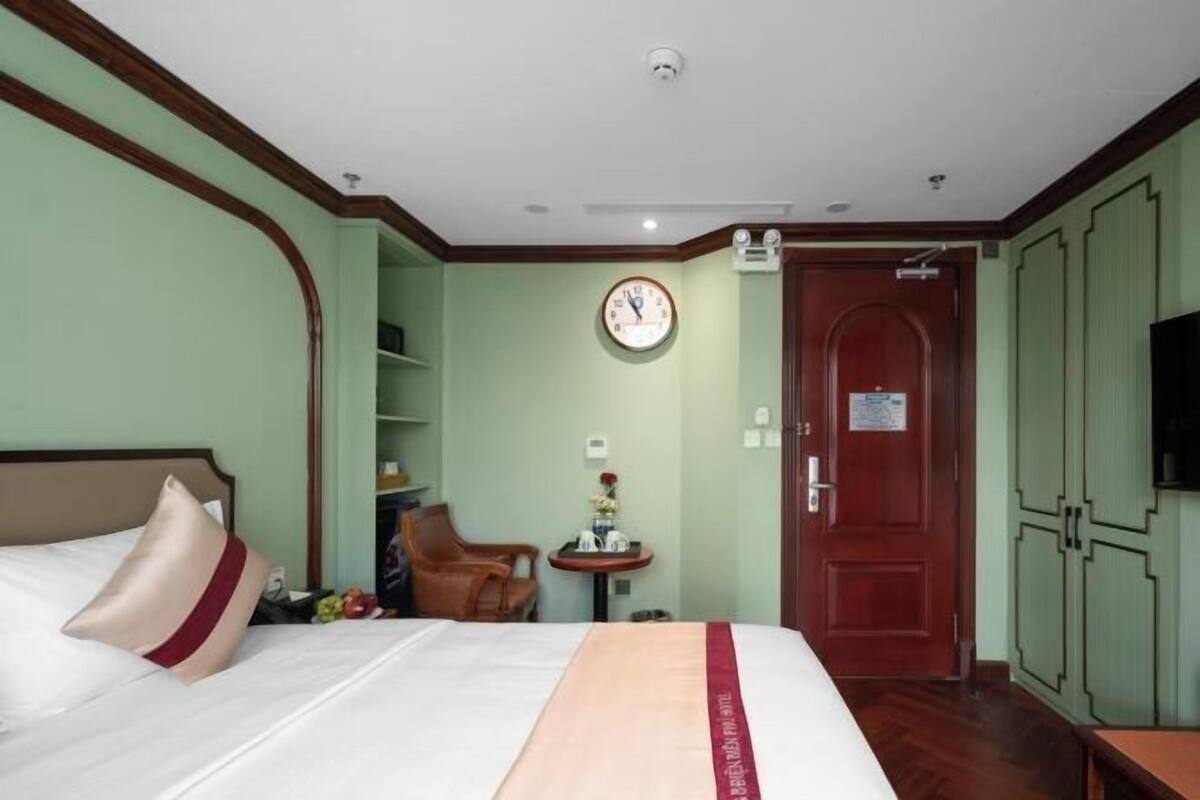 A hotel room with a bed, a clock, a door, and a small table with a vase.