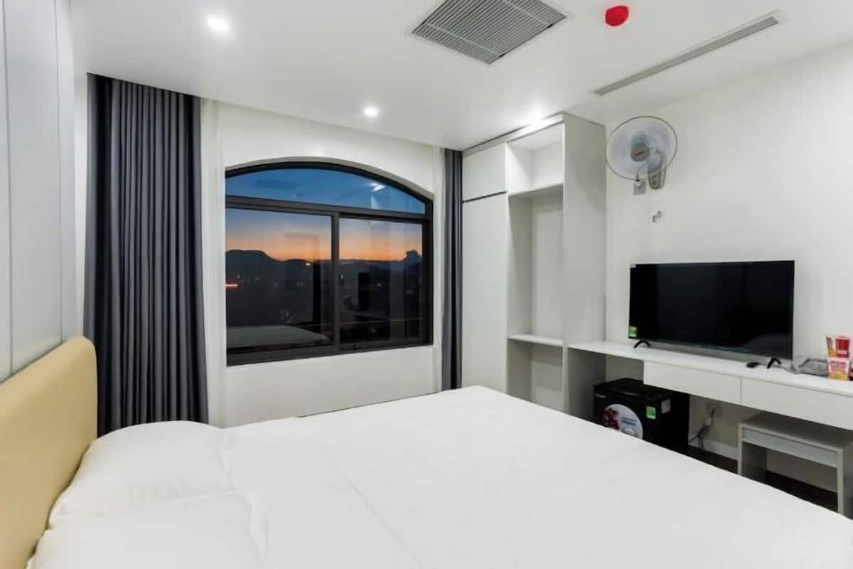 A hotel room with a bed, a TV, a desk, a window with a view, and a ceiling fan.