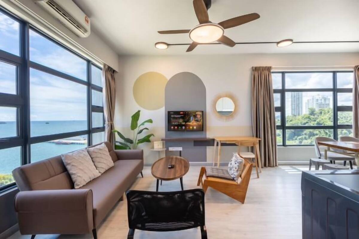 A modern living room with a sofa, coffee table, and dining area, large windows with a view of the sea, and a ceiling fan.