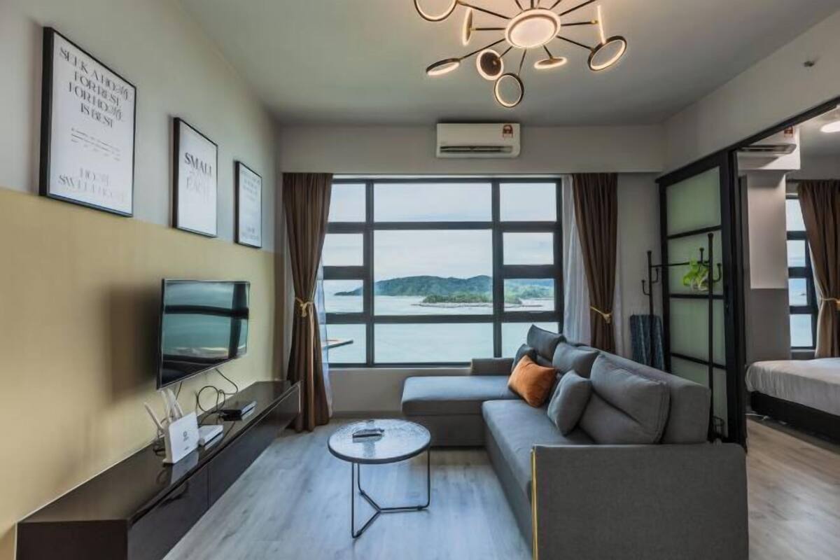 A modern hotel room with a large window offering a view of the sea, a sofa, a small table, and a flat-screen TV.