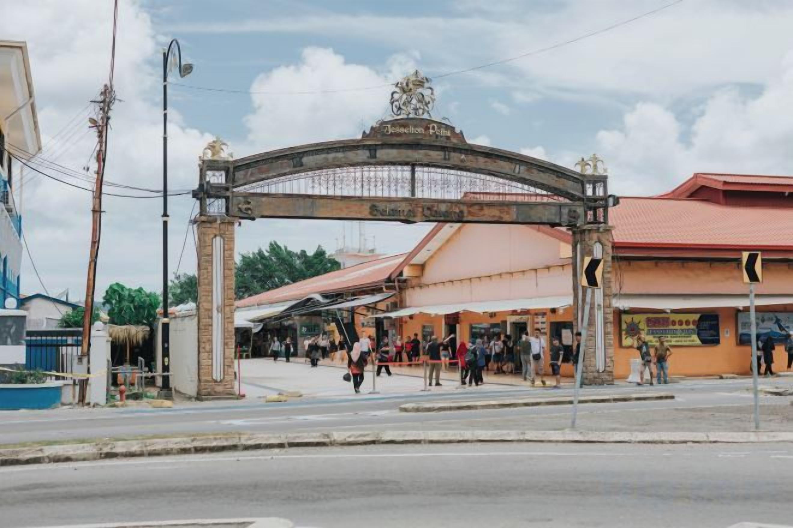 A street entrance with a large archway, a building with a red roof, and people walking by.