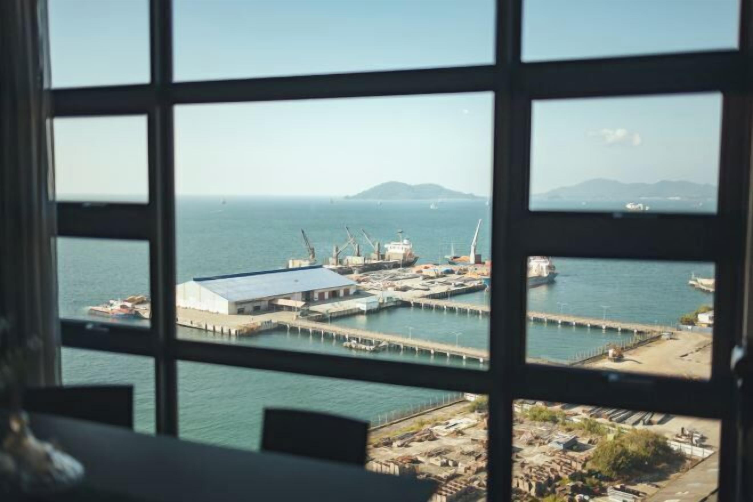 A view through a window of a coastal industrial area with a dock, cranes, and a large building.