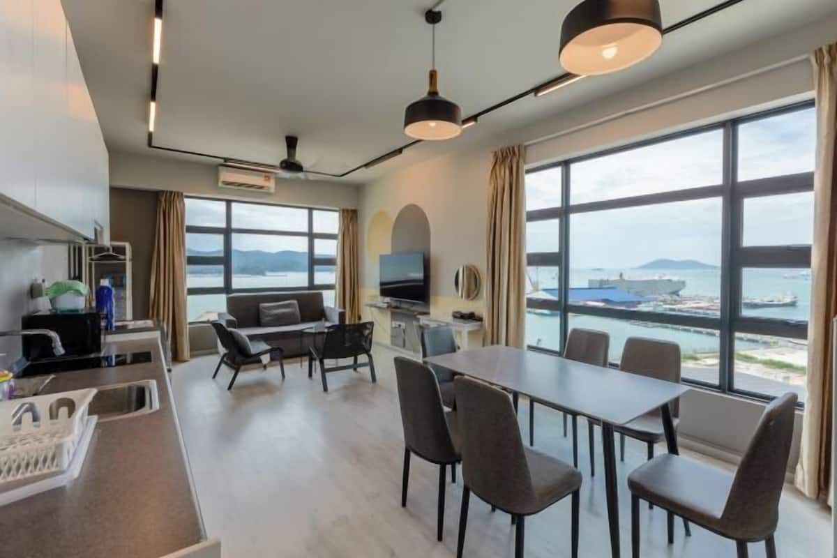 A modern hotel room with a dining area, a sofa, a TV, and large windows offering a view of the sea.