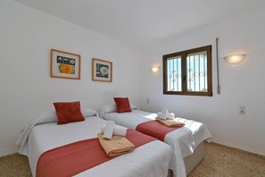 Quarto de hotel com duas camas, cada uma com almofada vermelha e edredom branco. Há quadros nas paredes, uma janela com vista e luminária fixada na parede.