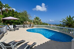 Uma piscina curva com espreguiçadeiras e um guarda-sol, rodeada por vegetação exuberante e um guarda-corpo branco.