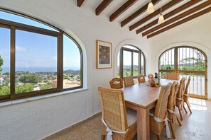 Uma sala de jantar com mesa de madeira e cadeiras de vime, grandes janelas em arco e vista para a paisagem.