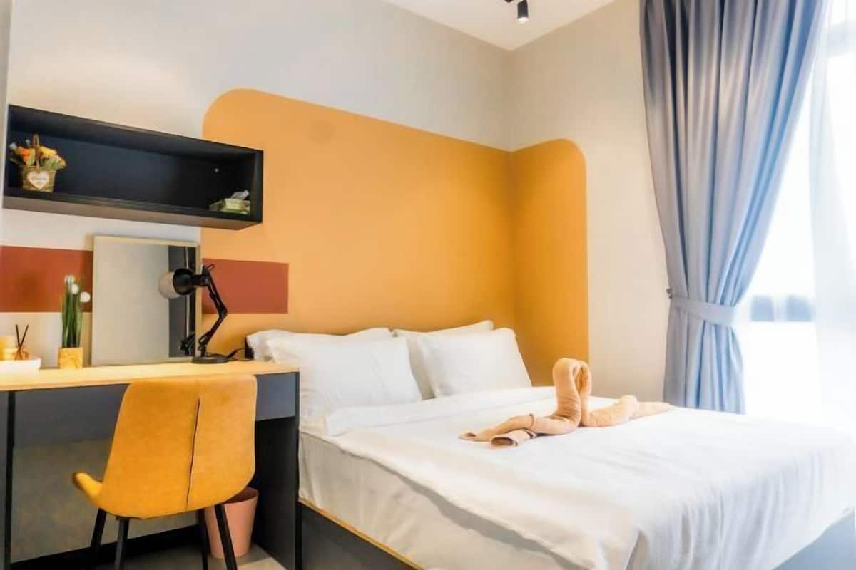 A hotel room with a bed, a desk with a chair, a mirror, and a shelf with items.