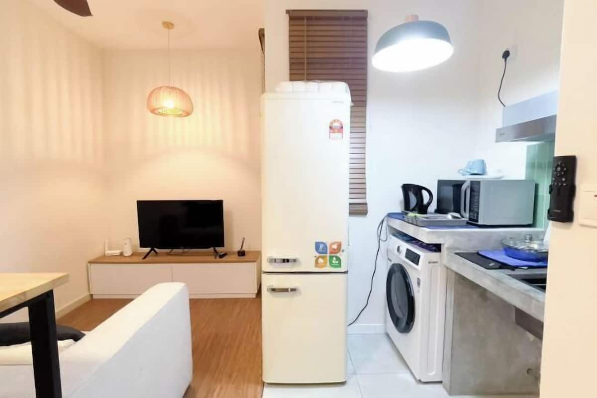 A compact living space with a flat-screen TV, a refrigerator with magnets, a washing machine, and a dining area with a table and chair.