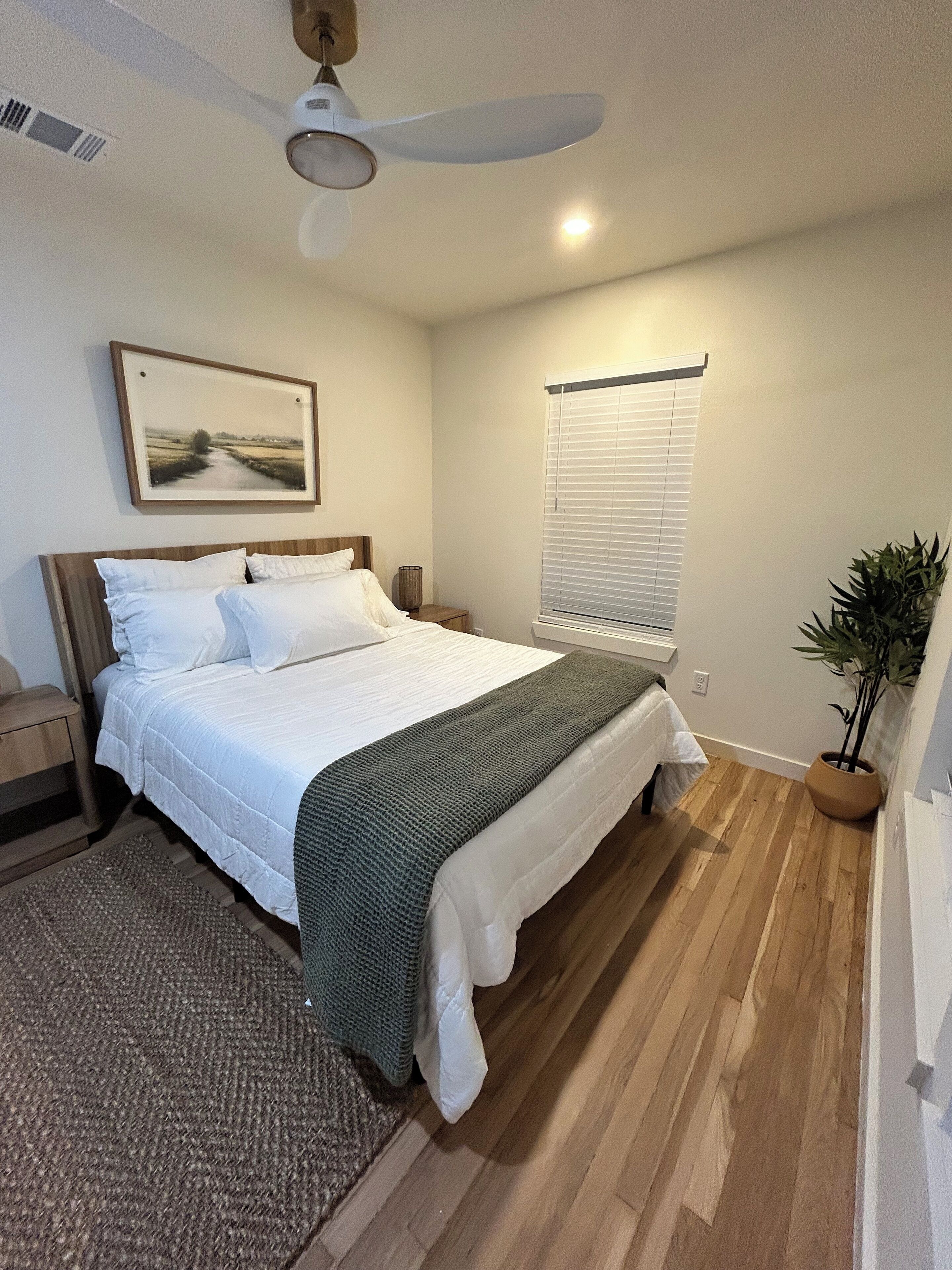 A bedroom with a bed, a nightstand, a ceiling fan, a window with blinds, and a potted plant.