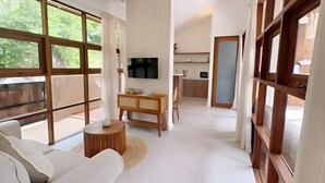 A spacious living area with a sofa, a wooden side table, a flat-screen TV mounted on the wall, and large windows offering a view of greenery outside. - Villa Lucky, Villa 13 (Koh Samui)