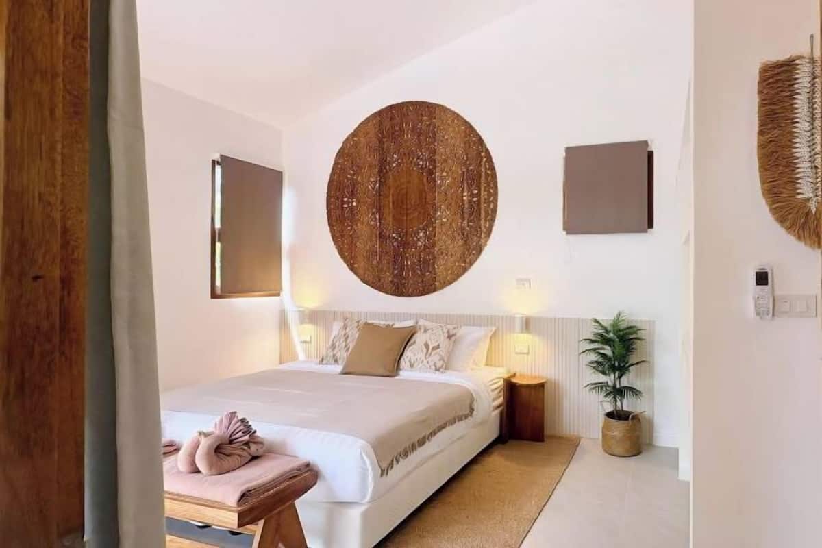 Quarto moderno com cama grande, mesas de cabeceira, uma obra de arte circular pendurada na parede e um vaso com planta.