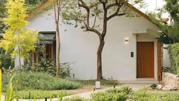 Un edificio blanco con puerta de madera, rodeado de un jardín con plantas diversas y un árbol.