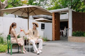 Two people seated at an outdoor table with a large umbrella, a brick wall, and a wooden gate in the background. - ดาวนับพัน Baan Phandao (Mae Rim)