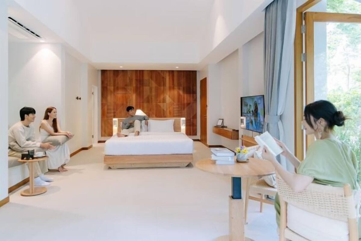 A modern hotel room with a large bed, a sitting area with a table, and a view of greenery outside.