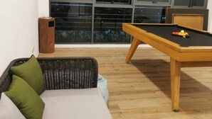 Quarto com mesa de bilhar, sofá de vime com almofadas e janela com vista para a cidade à noite.