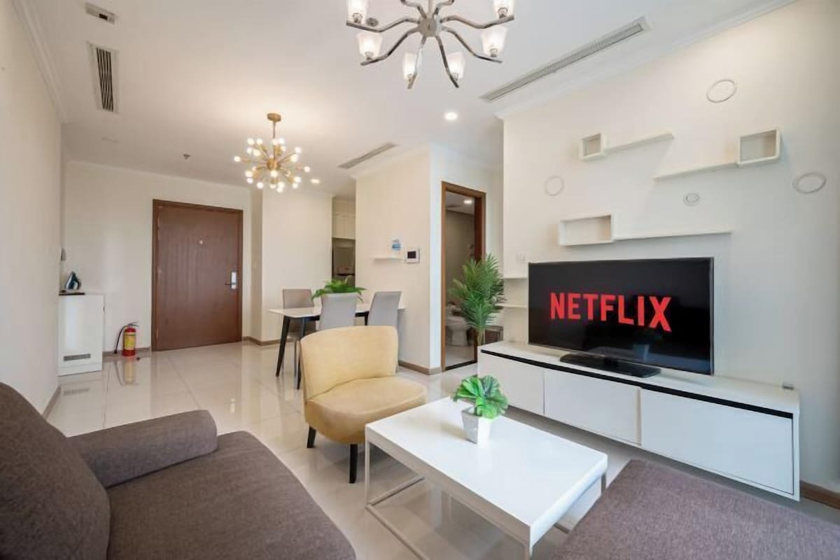 Un salon moderne avec un canapé, une table basse, un téléviseur affichant le logo « NETFLIX », un lustre et, au fond, un coin repas.