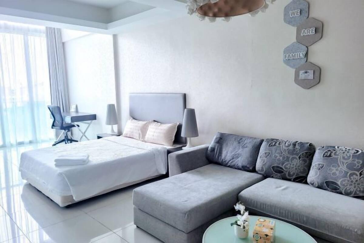 A modern hotel room with a large bed, a grey sofa, a small table with a vase, and a wall with hexagonal decor pieces.
