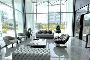 A modern living room with a glass wall, a grey sofa, a white tufted ottoman, and a black and white patterned chair. - Baysuites Near Sicc (Kota Kinabalu)