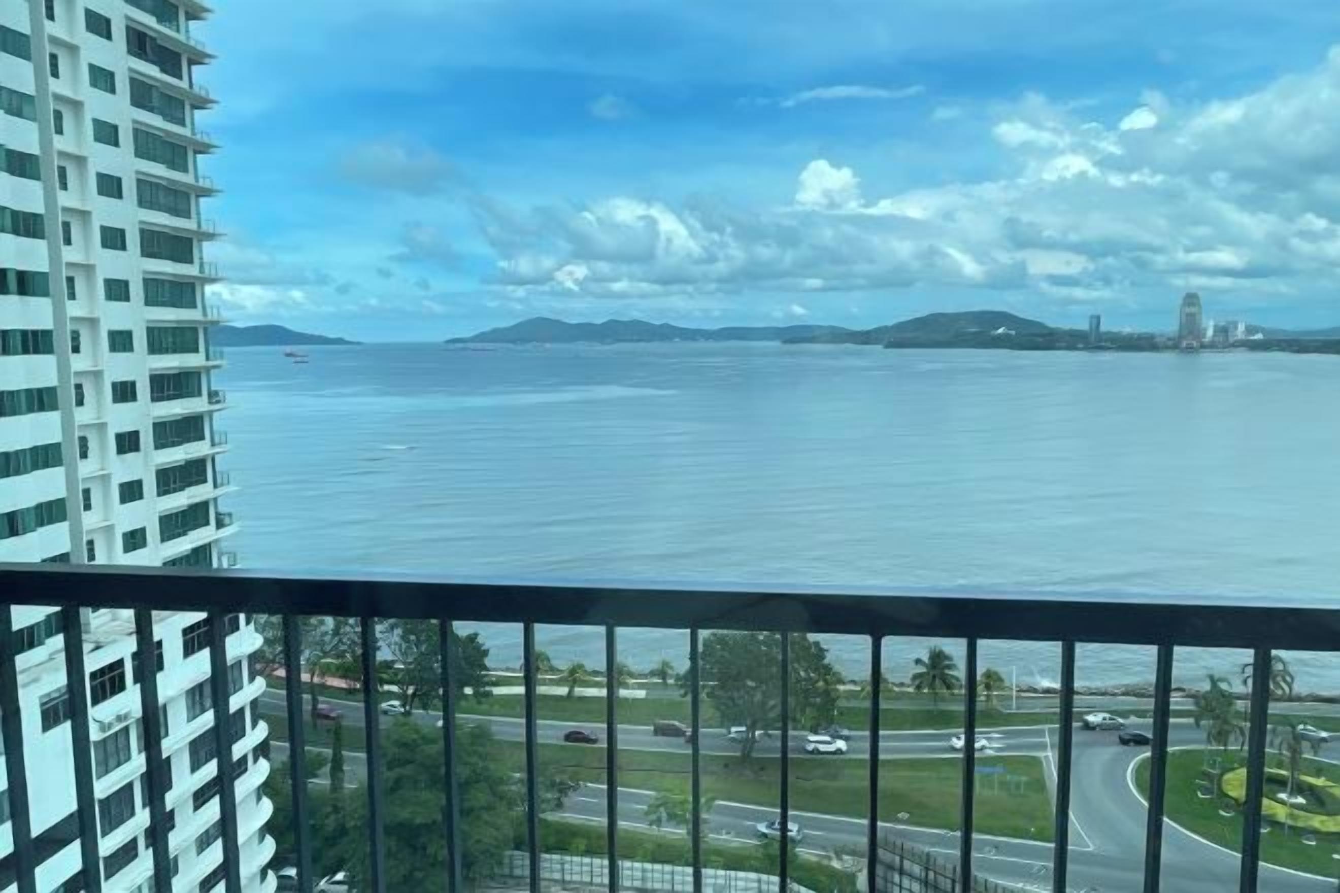 A view from a balcony overlooking a body of water, with a road and greenery in the foreground.