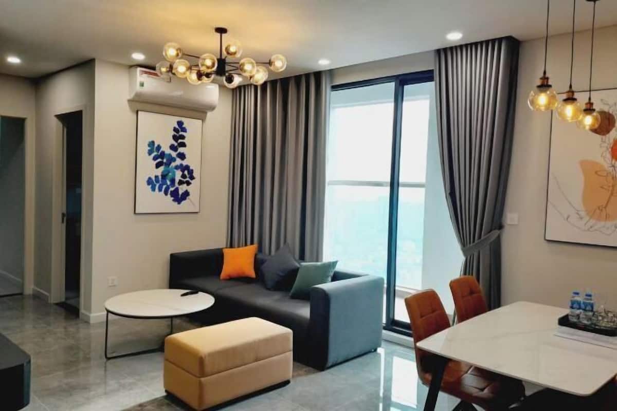 A modern living room with a sofa, ottoman, and dining table. There is a large window with curtains, a ceiling light fixture, and artwork on the wall.