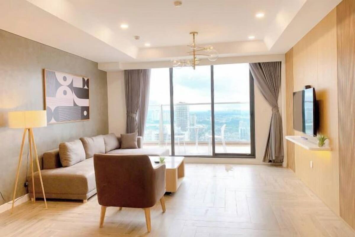 A modern living room with a sofa, armchair, and coffee table, large windows with curtains, a flat-screen TV, and a painting on the wall.