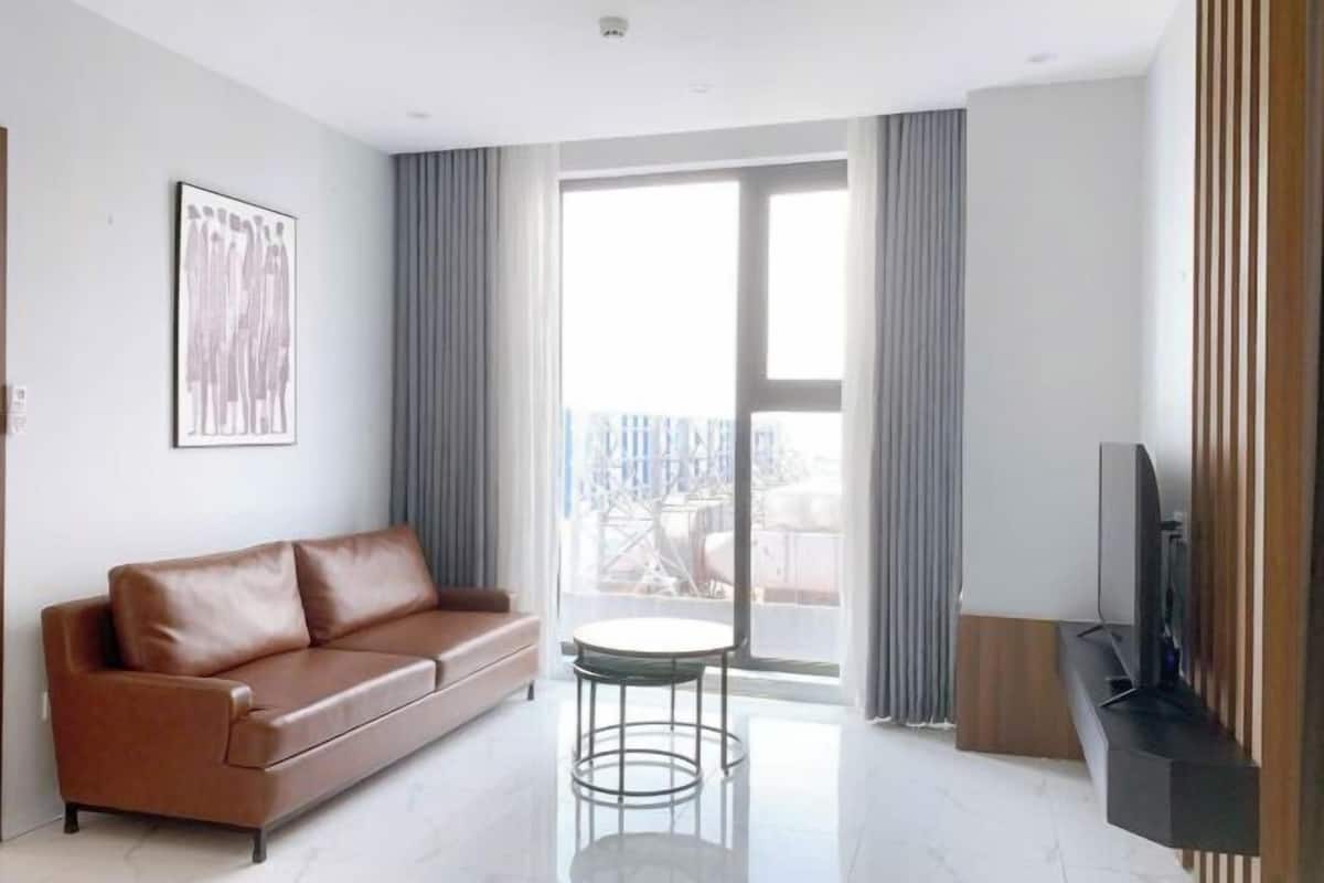 A modern living room with a brown leather sofa, a small round table, a flat-screen TV, and a large window with curtains.