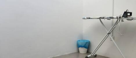 Esquina de una habitación con una silla plegable de metal, un cubo de basura azul y una pared blanca.