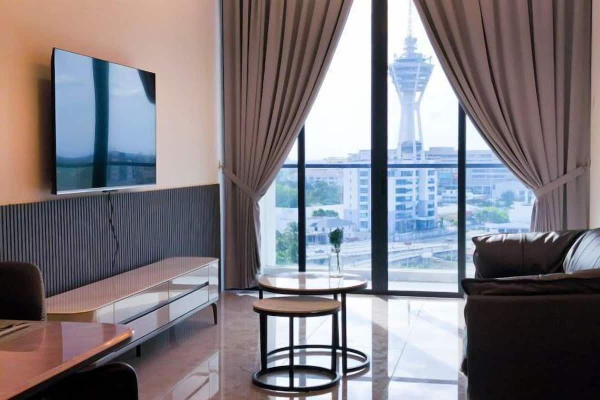 A modern hotel room with a large window, a flat-screen TV, a sofa, a small round table, and a bench.