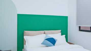 Una cama con ropa de cama blanca y un cojín de acento azul, una pared verde y una ventana con persianas.
