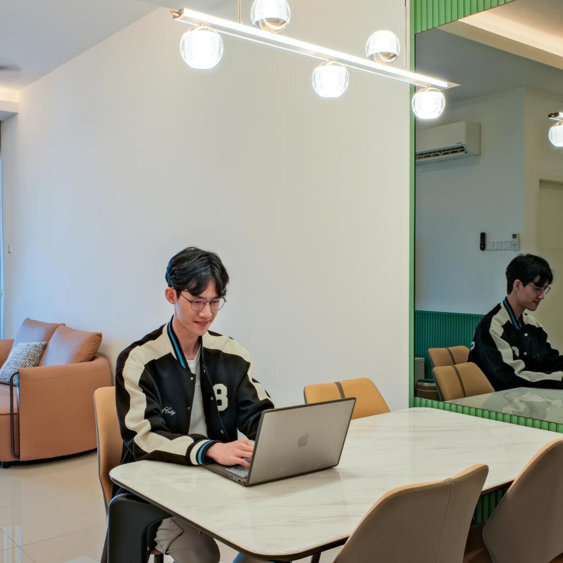 A person sitting at a table with a laptop in a room with a mirror, a couch, and a ceiling light fixture.
