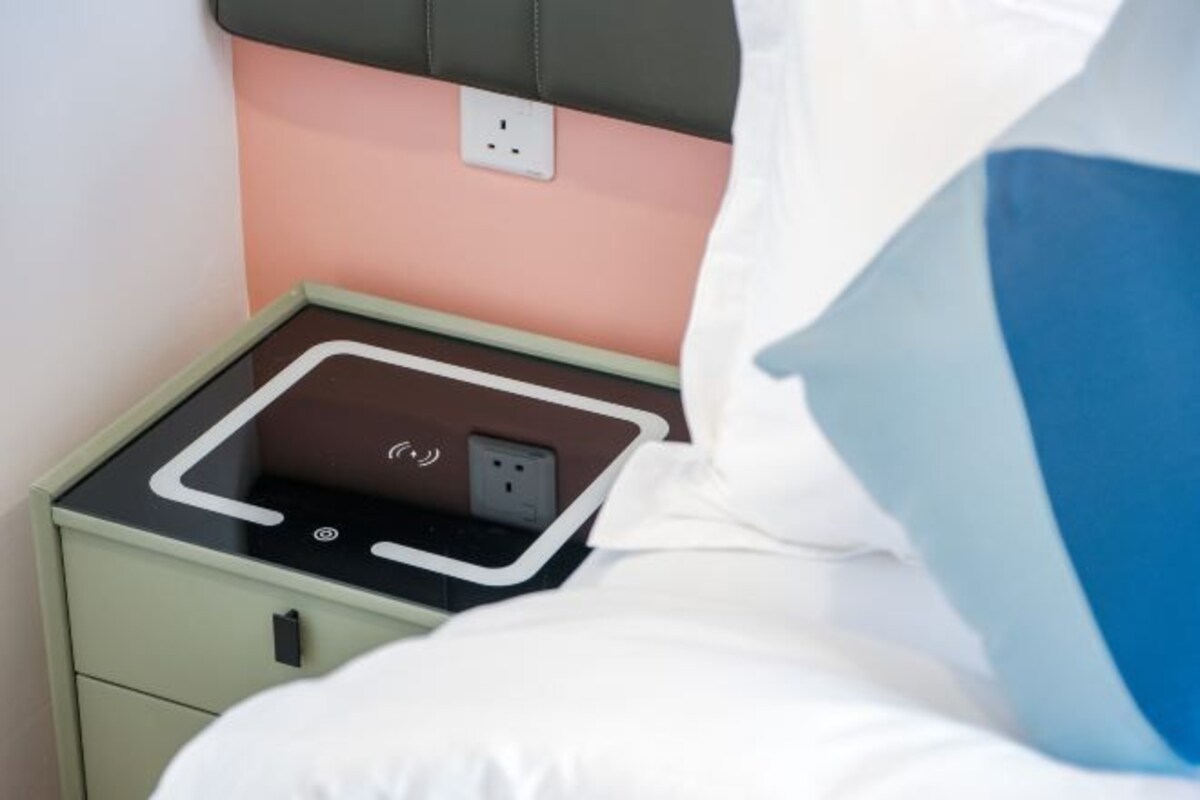 A bed with a white comforter, a green nightstand with a power outlet, and a pink wall.