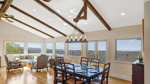 A spacious dining area with a large wooden table and chairs, a chandelier, and large windows offering a view of the landscape.