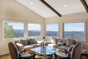 A spacious living room with a glass coffee table, beige sofas, and large windows offering a view of the mountains.