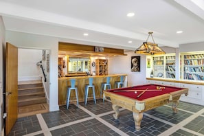 A pool table in a room with a bar area, bookshelves, and a staircase.