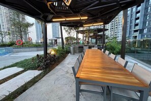 An outdoor seating area with a long wooden table and benches, surrounded by greenery and a city view. - LUMIERE RIVERSIDE CONDOTEL IN CENTER HO CHI MINH (Ho Chi Minh City)