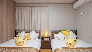 Une chambre d’hôtel avec deux lits, chacun agrémenté d’un oreiller à l’effigie de Pikachu, une table de chevet avec une lampe et un climatiseur fixé au mur.