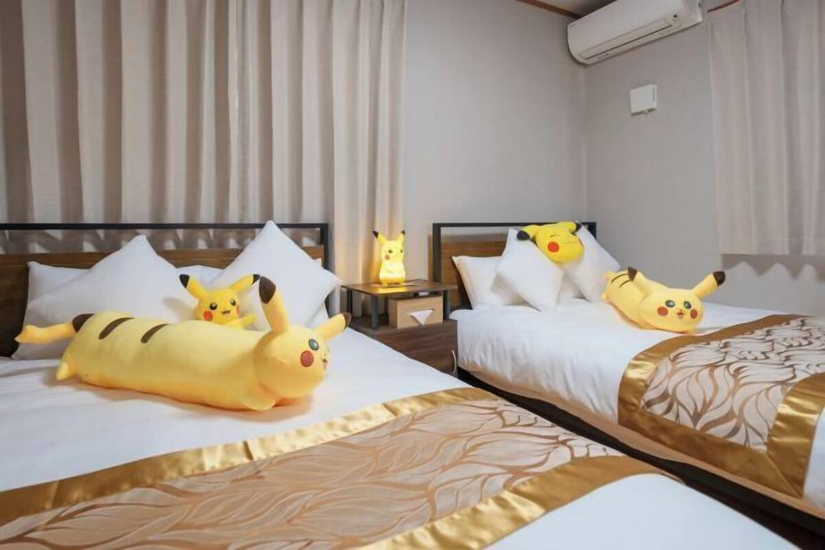 A hotel room with two beds, each with a Pikachu plush toy, white bedding, and a patterned gold accent.
