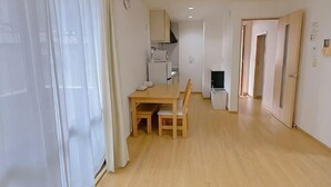 Habitació amb taula de dinar de fusta, cadires, una cuina petita i una porta que dona a una altra habitació.