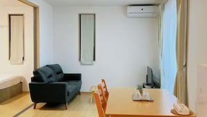 Una habitació amb un llit, un sofà negre, una taula de dinar de fusta i una televisió.