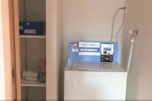 A white washing machine with a blue control panel, a blue box on a shelf, and a white wall with a power outlet. - Uenohouse Annex (Tokyo)