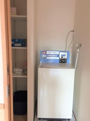 Un gabinete blanco con una lavadora COIN azul y blanca encima, un enchufe en la pared y una puerta parcialmente visible.