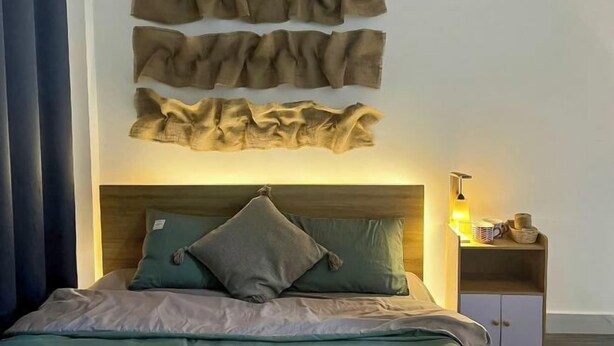 A bed with a wooden headboard, a green pillow, and a beige blanket. There is a nightstand with a lamp and a cup beside the bed. - Small Private house in Bui Vien Walking Street (Ho Chi Minh City)