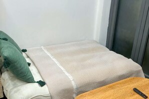 En liten, välordnad säng med ett beige täcke och gröna kuddar, ett träbord med en fjärrkontroll och ett fönster med persienner.