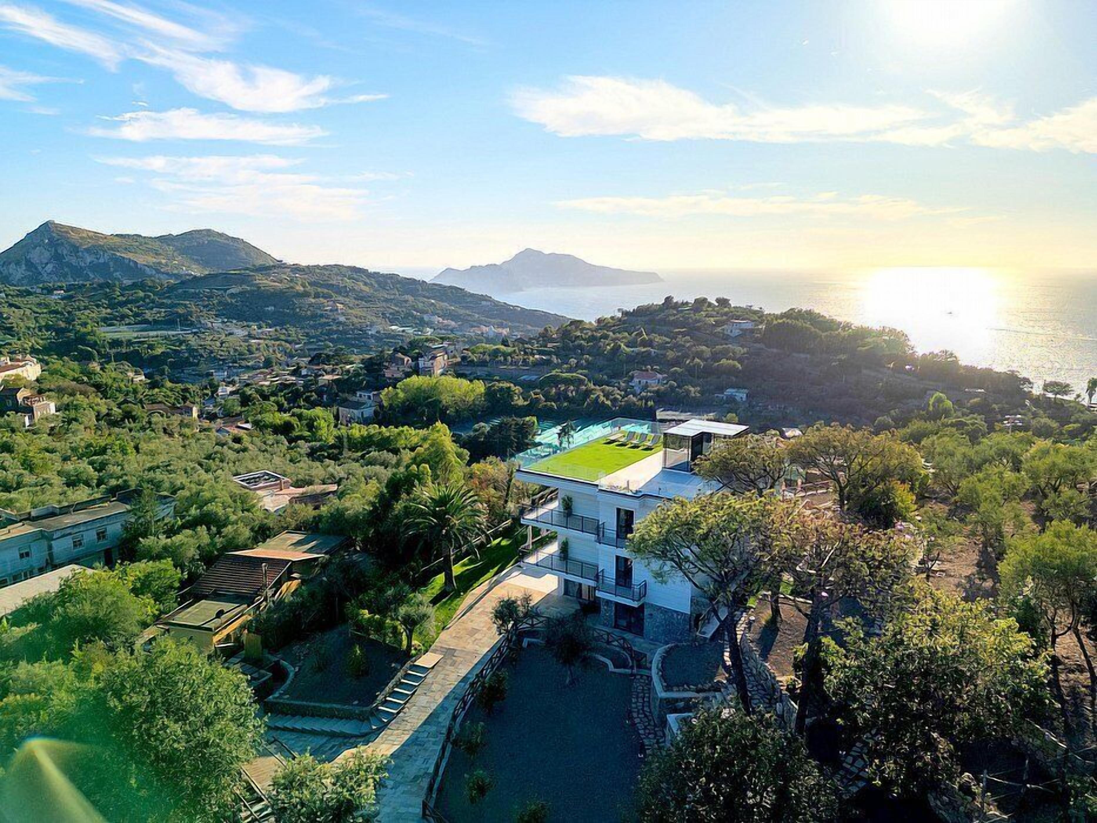 A modern villa with a rooftop terrace, surrounded by lush greenery and overlooking a coastal landscape with mountains in the distance.