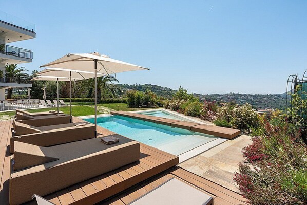 A swimming pool with sun loungers and umbrellas, surrounded by a landscaped garden and a view of the hills.