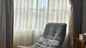 A room with a large window covered by sheer curtains, a dark leather armchair, and a small wooden side table with a lamp. - LYLAC Home Saigon HHH - Beautiful City Center Whole Townhouse in D1 (Ho Chi Minh City)