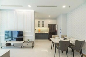 A modern living room with a flat-screen TV, a dining table with chairs, and a kitchen area in the background. - Saigon Landmark Apartment - Vinhomes (Ho Chi Minh City)