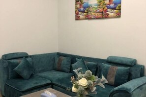 A teal sectional sofa with multiple cushions, a coffee table with a vase of flowers, and a colorful landscape painting on the wall. - Haven Inn (Phnom Penh)
