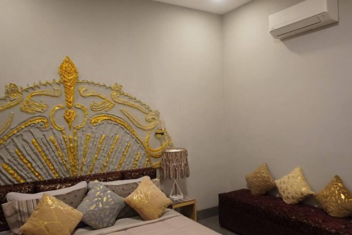 A bedroom with a decorative headboard, a bed with multiple pillows, a side table with a lamp, and a dark-colored sofa.