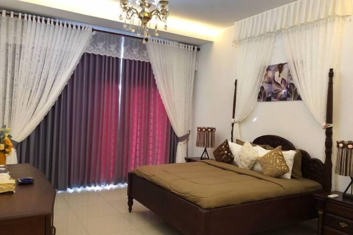 A bedroom with a bed, curtains, a chandelier, a bedside table, and a wall-mounted lamp.